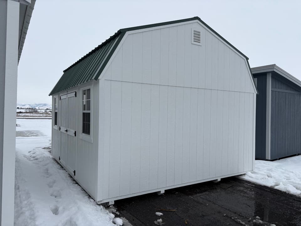 White 14x16 side lofted barn with a hunter green gambrel roof and gable vent, standing in the snow on the JTCS Buildings lot